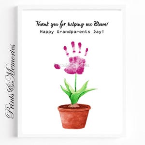 Op de afbeelding: Een ingelijste print met een aquarel illustratie van een bloem gemaakt van een handafdruk. De roze bloem heeft groene bladeren en een bruine pot. De tekst luidt: "Bedankt dat je me helpt bloeien! Fijne grootoudersdag!"
