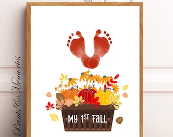 May include: A white background with a pair of red baby footprints in the shape of a heart. Below the footprints is a brown basket filled with orange, yellow, and red pumpkins, fall leaves, and flowers. The basket has the text "MY 1ST FALL" on the front.
