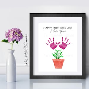 May include: A framed print with a black border features a watercolor illustration for Mother's Day. The artwork includes pink handprints forming a flower, a terracotta pot, and the text "Happy Mother's Day, I love you!" A white vase holds a purple flower.