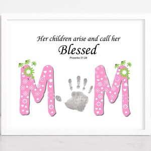 Op de afbeelding: Een ingelijste print met het woord "Blessed" in sierlijke letters. Het woord "MOM" is geschreven in roze letters versierd met witte bloemen en hartjes. Een grijze handafdruk staat in het midden. De tekst "Her children arise and call her" staat erboven.
