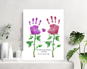 May include: Watercolor print featuring two handprint flowers in shades of purple and red. Green stems and leaves support the flowers. The text reads "Thank you for helping us grow! Made with love by".