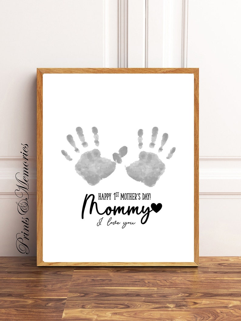 Pu&ograve; includere: Una stampa d'arte stampabile bianca con due impronte di mani grigie e il testo "Happy 1st Mother's Day! Mommy I love you" in nero con un cuore.