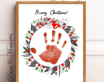 Puede incluir: Una impresión enmarcada con una huella de mano dentro de una corona. La corona presenta detalles en gris, verde y rojo, con las palabras "Merry Christmas!" en la parte superior. El marco es de color marrón claro.