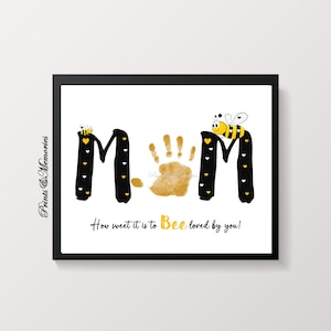 Op de afbeelding: Een zwart-witte printbare kunstprint met het woord "MOM" in grote letters. De letter "O" is een handafdruk in gouden verf. Er zijn kleine gele bijen en harten rond de letters. De tekst "How sweet it is to Bee loved by you!" staat onder de letters.