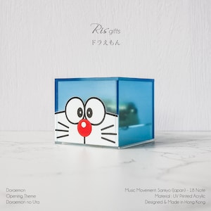 May include: A blue acrylic music box featuring the cartoon character Doraemon. The box has a cartoon face design with large white eyes, a red nose, and white whiskers. The text "Ris gifts" and Japanese characters are visible.