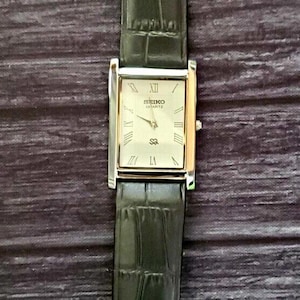 May include: A rectangular Seiko Quartz watch with a silver and gold-toned case and a black leather strap. The watch face is cream-coloured with Roman numerals and gold-toned hands. The strap has a textured, crocodile-like pattern.