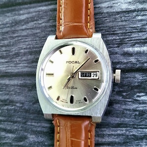 May include: A vintage Focal De Luxe wristwatch with a silver dial and a brown leather strap. The watch face displays the day and date, with the text "5-FRI 29". The watch has a silver-coloured case and a brown leather band.