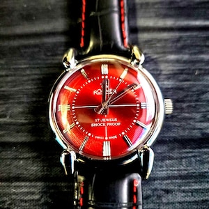 Puede incluir: Un reloj de pulsera con esfera roja y correa de cuero negro con costuras rojas. La esfera del reloj tiene agujas y marcadores plateados, y el texto "17 JEWELS SHOCK PROOF" y "SWISS MADE".
