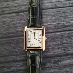 Puede incluir: Un reloj de pulsera rectangular con una caja dorada y una esfera blanca con números romanos negros. El reloj tiene una correa de cuero negro con un patrón de cocodrilo. La esfera del reloj muestra la marca "PARI RAFZ".