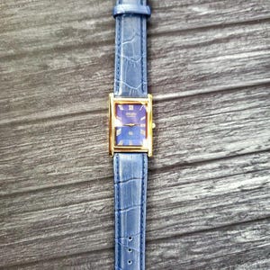 May include: A gold-tone Seiko watch with a blue face and a blue leather band. The watch has a rectangular face and Roman numeral hour markers.