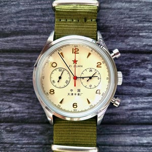 May include: A classic wristwatch with a cream dial, red star, and the text "21 ZUAN". The watch has a silver case and a green nylon strap. The watch face displays sub-dials and the text "中国 天津手表厂". The strap is olive green.