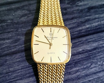 May include: A gold-toned quartz wristwatch with a square face and a woven gold band. The watch face is cream-colored with black markers and hands. The watch face has the text "Quartz" and the brand name "Lars Christensen Since 1950".