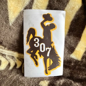 May include: A sticker featuring a brown silhouette of a cowboy riding a bucking bronco, with the number "307" in white. The design has a yellow outline and is on a white background.