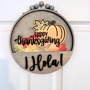 Puede incluir: Un letrero de pared circular de madera con un dise&ntilde;o en capas que presenta una calabaza de color naranja claro, hojas de oto&ntilde;o coloridas y el texto "HAPPY thanksgiving" en negro. Debajo, la palabra "&iexcl;Hola!" aparece en negro, con cuentas grises.