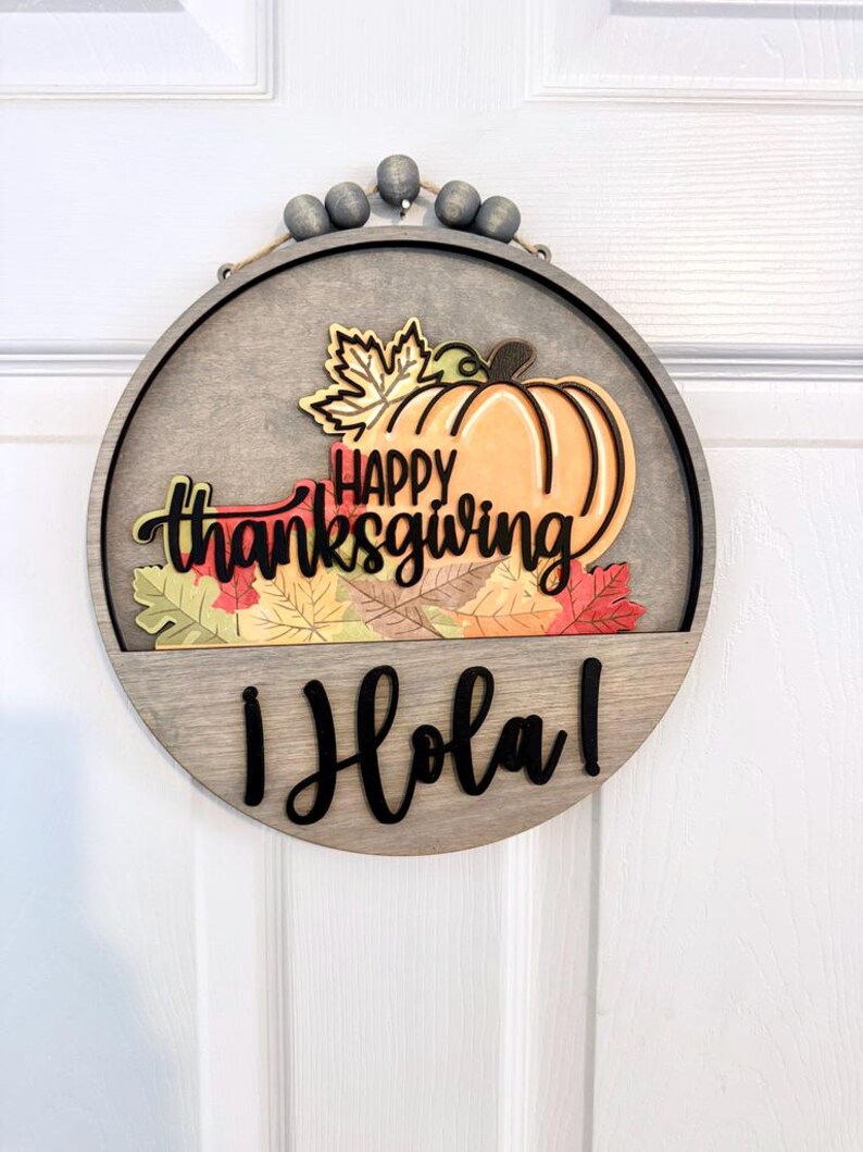 Puede incluir: Un letrero de pared circular de madera con un dise&ntilde;o en capas que presenta una calabaza de color naranja claro, hojas de oto&ntilde;o coloridas y el texto "HAPPY thanksgiving" en negro. Debajo, la palabra "&iexcl;Hola!" aparece en negro, con cuentas grises.