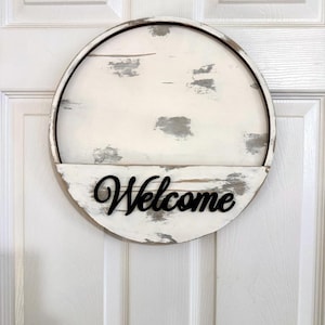 Puede incluir: Un letrero circular de madera con un acabado desgastado en blanco y gris. El letrero tiene un estante horizontal en la mitad inferior con la palabra Welcome escrita en letras cursivas negras en relieve fijadas en la superficie.