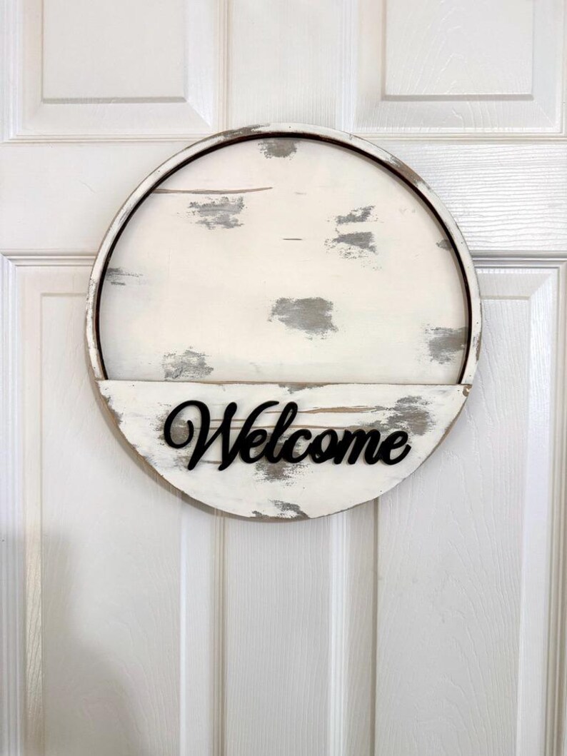 Puede incluir: Un letrero circular de madera con un acabado desgastado en blanco y gris. El letrero tiene un estante horizontal en la mitad inferior con la palabra Welcome escrita en letras cursivas negras en relieve fijadas en la superficie.
