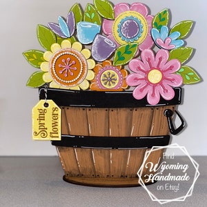 May include: Wooden spring flowers in a brown bucket with a black rim and handle. The flowers are pink, yellow, blue, and purple with green leaves. A yellow tag reads "Spring flowers".
