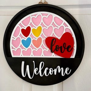 Puede incluir: Un letrero de pared circular de madera negra con un recorte arqueado blanco lleno de corazones rosas. Un coraz&oacute;n rojo grande con la palabra love en cursiva negra est&aacute; fijado a la derecha. La palabra Welcome est&aacute; escrita en blanco en la base.