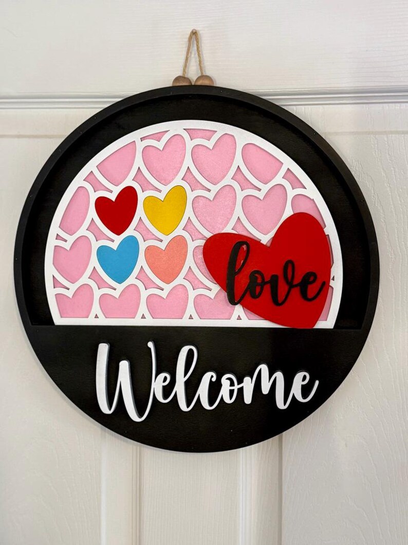Puede incluir: Un letrero de pared circular de madera negra con un recorte arqueado blanco lleno de corazones rosas. Un coraz&oacute;n rojo grande con la palabra love en cursiva negra est&aacute; fijado a la derecha. La palabra Welcome est&aacute; escrita en blanco en la base.