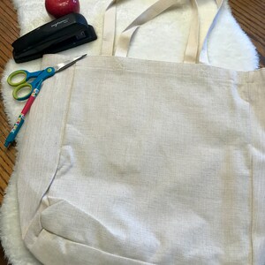 May include: A white canvas tote bag with two long straps. The bag is lying flat on a white fluffy surface. A red apple, a stapler, and a pair of scissors are visible in the background.