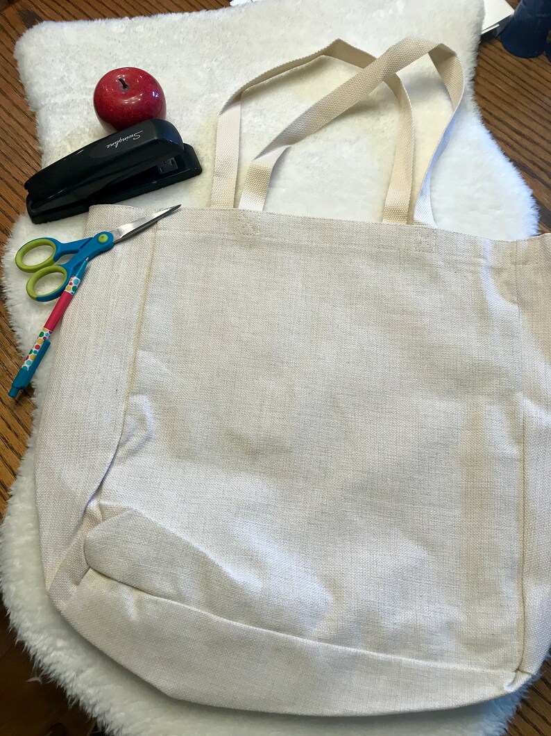 May include: A white canvas tote bag with two long straps. The bag is lying flat on a white fluffy surface. A red apple, a stapler, and a pair of scissors are visible in the background.