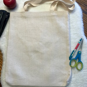 May include: A plain white canvas tote bag with a long, cream-colored strap. The bag is lying flat on a white fluffy surface.