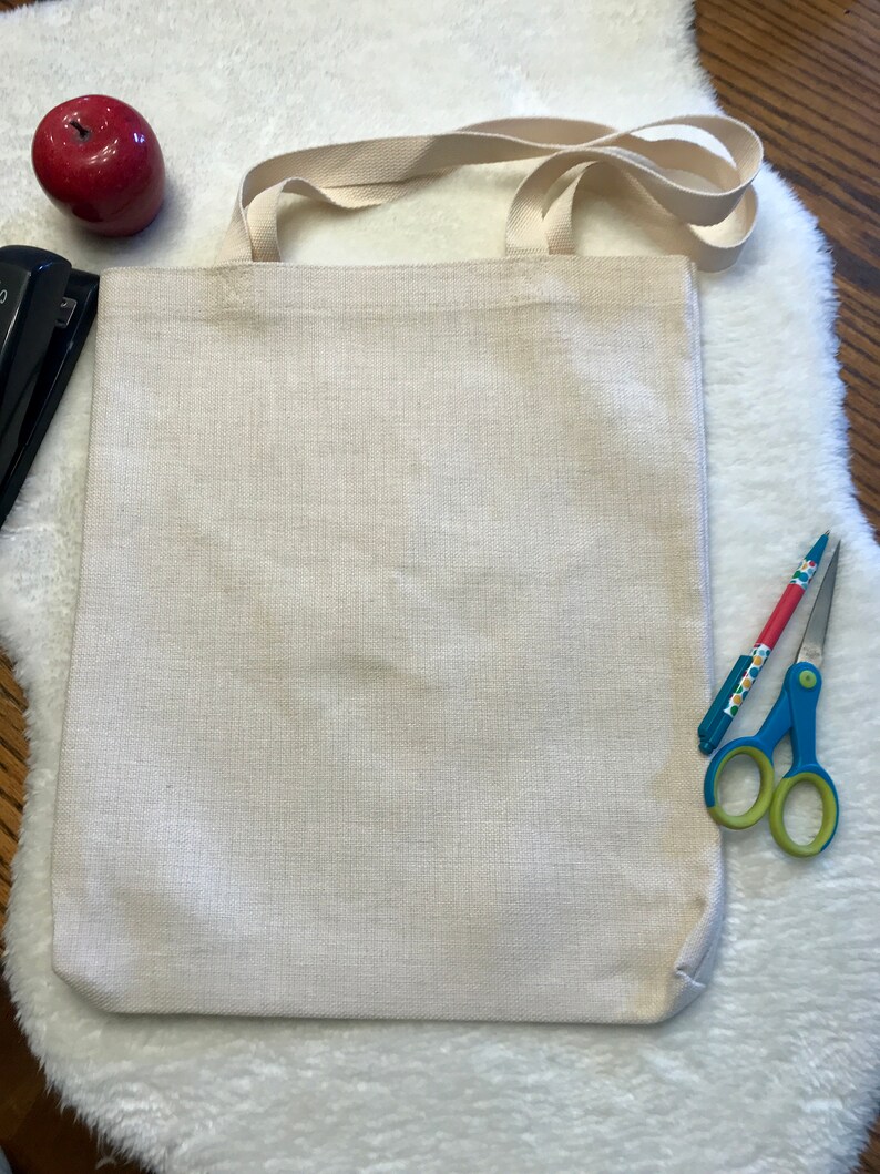 May include: A plain white canvas tote bag with a long, cream-colored strap. The bag is lying flat on a white fluffy surface.