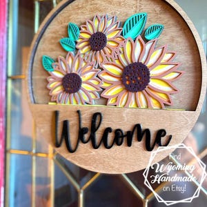 Puede incluir: Un letrero de puerta circular de madera con tres girasoles superpuestos de p&eacute;talos amarillos y centros marr&oacute;n oscuro, acentuados por hojas color verde azulado. La palabra Welcome est&aacute; escrita en letras cursivas negras en la parte inferior.