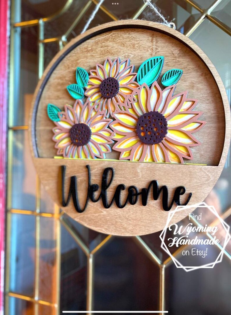 Puede incluir: Un letrero de puerta circular de madera con tres girasoles superpuestos de p&eacute;talos amarillos y centros marr&oacute;n oscuro, acentuados por hojas color verde azulado. La palabra Welcome est&aacute; escrita en letras cursivas negras en la parte inferior.