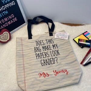May include: A beige canvas tote bag with black handles and a red and white striped design. The bag has the text "Does this bag make my papers look graded?" and "Mrs. Young" printed on it.