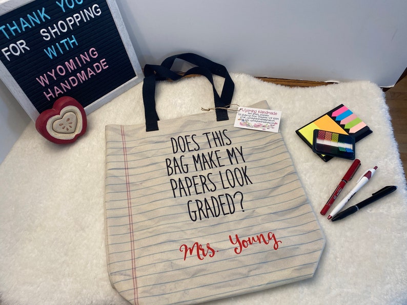 May include: A beige canvas tote bag with black handles and a red and white striped design. The bag has the text "Does this bag make my papers look graded?" and "Mrs. Young" printed on it.
