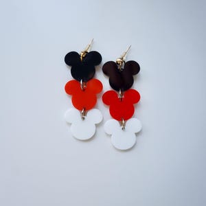 May include: A pair of dangle earrings featuring three stacked Mickey Mouse head shapes. The top shape is black, the middle shape is red, and the bottom shape is white.