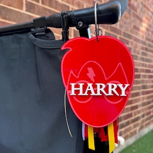 Puede incluir: Llavero de acrílico rojo con un rayo y el nombre "HARRY" en letras blancas. El llavero está sujeto a un asa de cochecito negro con cintas amarillas, rojas y blancas.