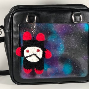 Pode incluir: Bolsa de ombro preta de couro sintético com uma janela transparente na frente. A janela apresenta uma estampa de galáxia e um brinquedo de pelúcia de crochê vermelho e preto pendurado em uma corrente prateada.
