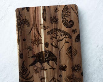 May include: A wooden cutting board with a dark brown wood grain and a black and white design of a crow, ferns, flowers, and leaves.