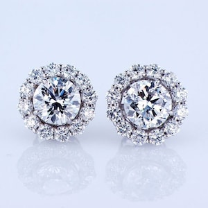 May include: A pair of diamond earrings with a halo setting. The earrings are made of white gold and feature a round brilliant cut diamond in the centre.