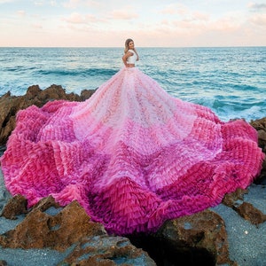 May include: A woman wearing a white top and a pink and white ombre ruffled dress stands on a rocky beach with the ocean in the background.