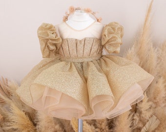 May include: A gold glitter tulle dress with puffed sleeves and a layered skirt. The dress is on a white mannequin and is surrounded by dried pampas grass.