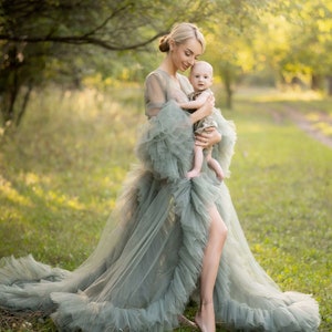 May include: A woman in a long, flowing, sage green tulle gown holds a baby in her arms. The woman and baby are standing in a grassy field with trees in the background.