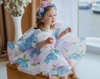 Op de afbeelding: Een jong meisje in een witte jurk met blauwe en roze bloemenprint, een bloemenkroon en gouden schoenen zit op een gele kubus.