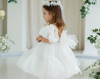 Può includere: Una bambina che indossa un abito bianco di tulle con decorazioni di perle e una corona di fiori bianchi. È in piedi su una superficie bianca con fiori bianchi sullo sfondo.