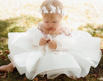 May include: A young child wearing a white dress with a flower crown and holding a small flower. The dress has a ruffled skirt and a sheer bodice with floral embroidery. The child is sitting on a grassy surface.