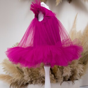 Peut inclure: Une robe en tulle fuchsia avec un col &agrave; volants et une jupe ample. La robe est expos&eacute;e sur un mannequin blanc.