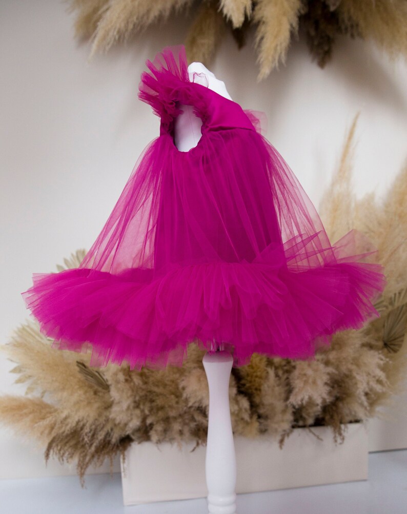 Peut inclure: Une robe en tulle fuchsia avec un col &agrave; volants et une jupe ample. La robe est expos&eacute;e sur un mannequin blanc.