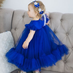 Peut inclure: Une jeune fille portant une robe en tulle bleu royal avec un bandeau de papillon blanc et bleu. La robe a un ourlet &agrave; volants et la fille est debout sur un canap&eacute; gris clair.