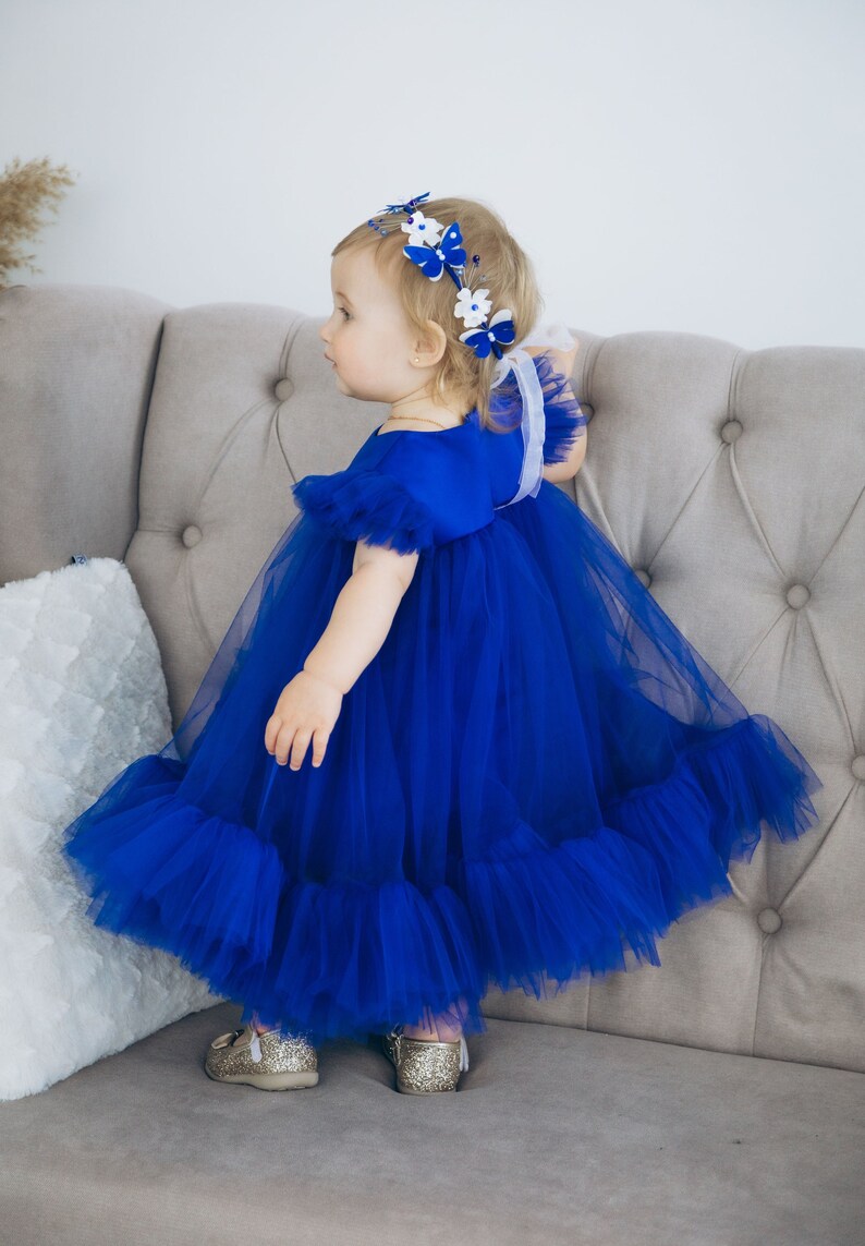 Peut inclure: Une jeune fille portant une robe en tulle bleu royal avec un bandeau de papillon blanc et bleu. La robe a un ourlet &agrave; volants et la fille est debout sur un canap&eacute; gris clair.