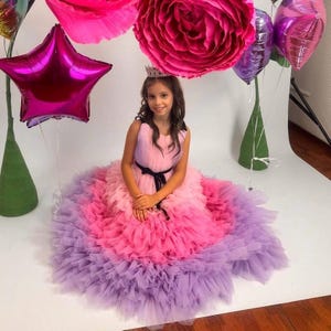 Könnte beinhalten: Eine junge Person trägt ein rosa, mehrschichtiges Kleid mit einem schwarzen Gürtel und einer kleinen Krone. Das Kleid wechselt von Rosa zu Lila. Der Hintergrund umfasst große rosa Papierblumen und sternförmige Ballons.