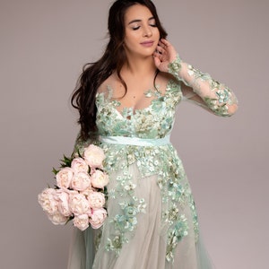 Op de afbeelding: Een vrouw in een lichtgroene, met bloemen geborduurde jurk met een transparante overlay, die een boeket roze pioenen vasthoudt.