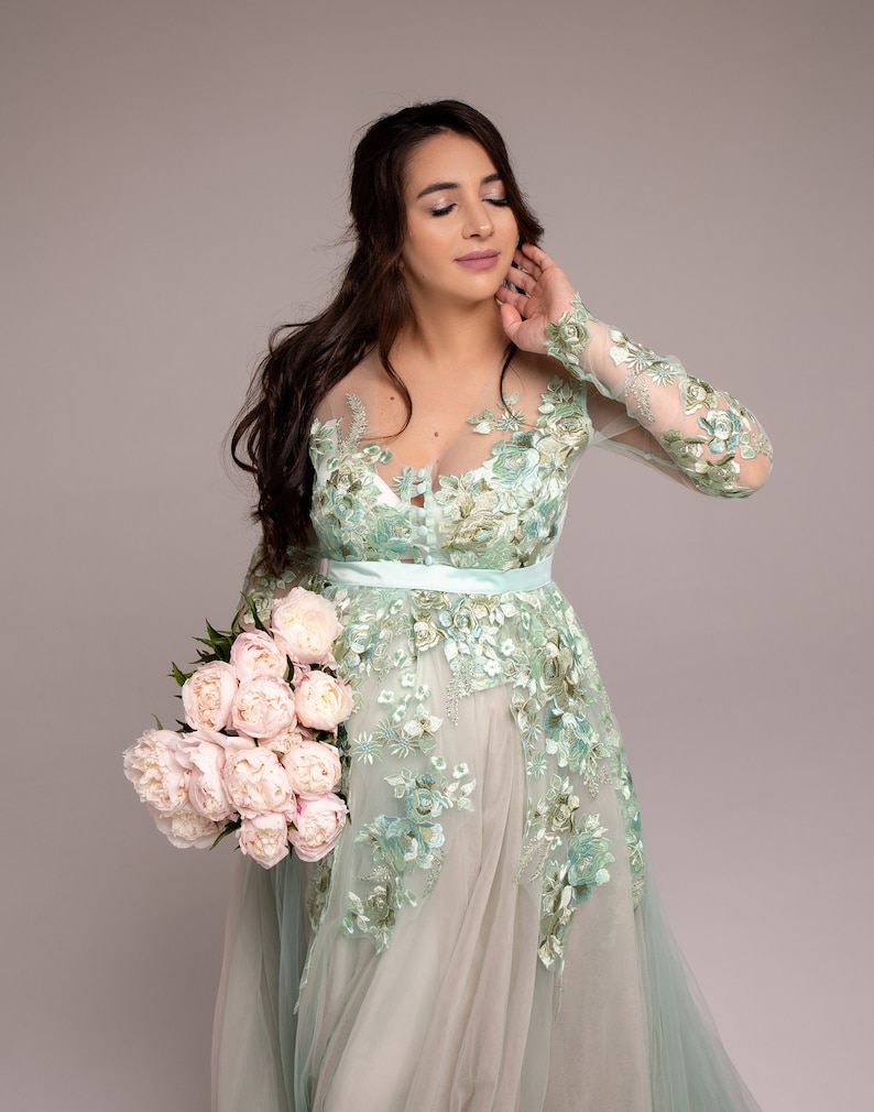 Op de afbeelding: Een vrouw in een lichtgroene, met bloemen geborduurde jurk met een transparante overlay, die een boeket roze pioenen vasthoudt.